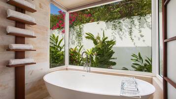 A modern bathroom with a freestanding bathtub, a wooden towel rack, and a large window with a view of lush greenery and flowers.
