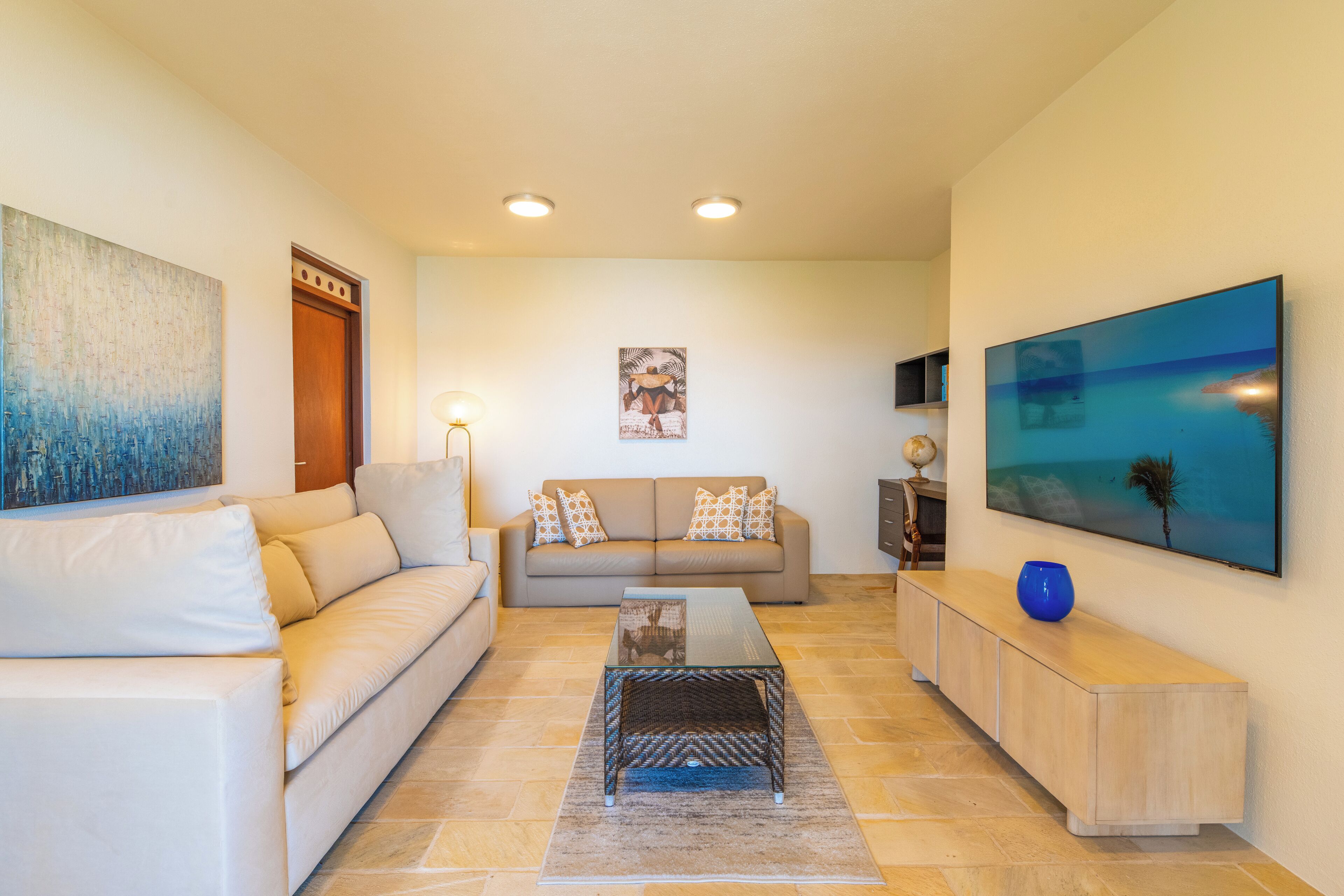 A living room with a beige sofa, a glass coffee table, a blue vase, and a large painting on the wall.