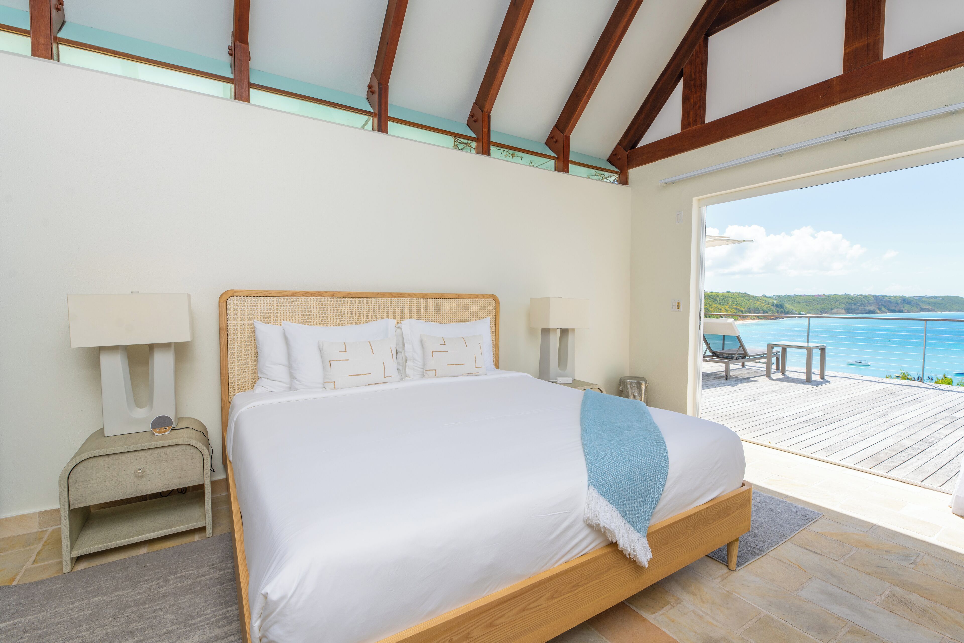 A bedroom with a large bed, bedside lamps, a nightstand, and a view of a balcony with outdoor seating.