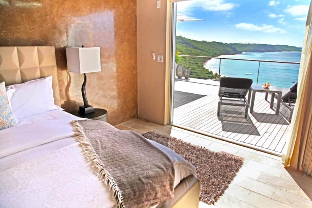 A bedroom with a large bed, a nightstand with a lamp, and a view of a balcony with a chair and table.