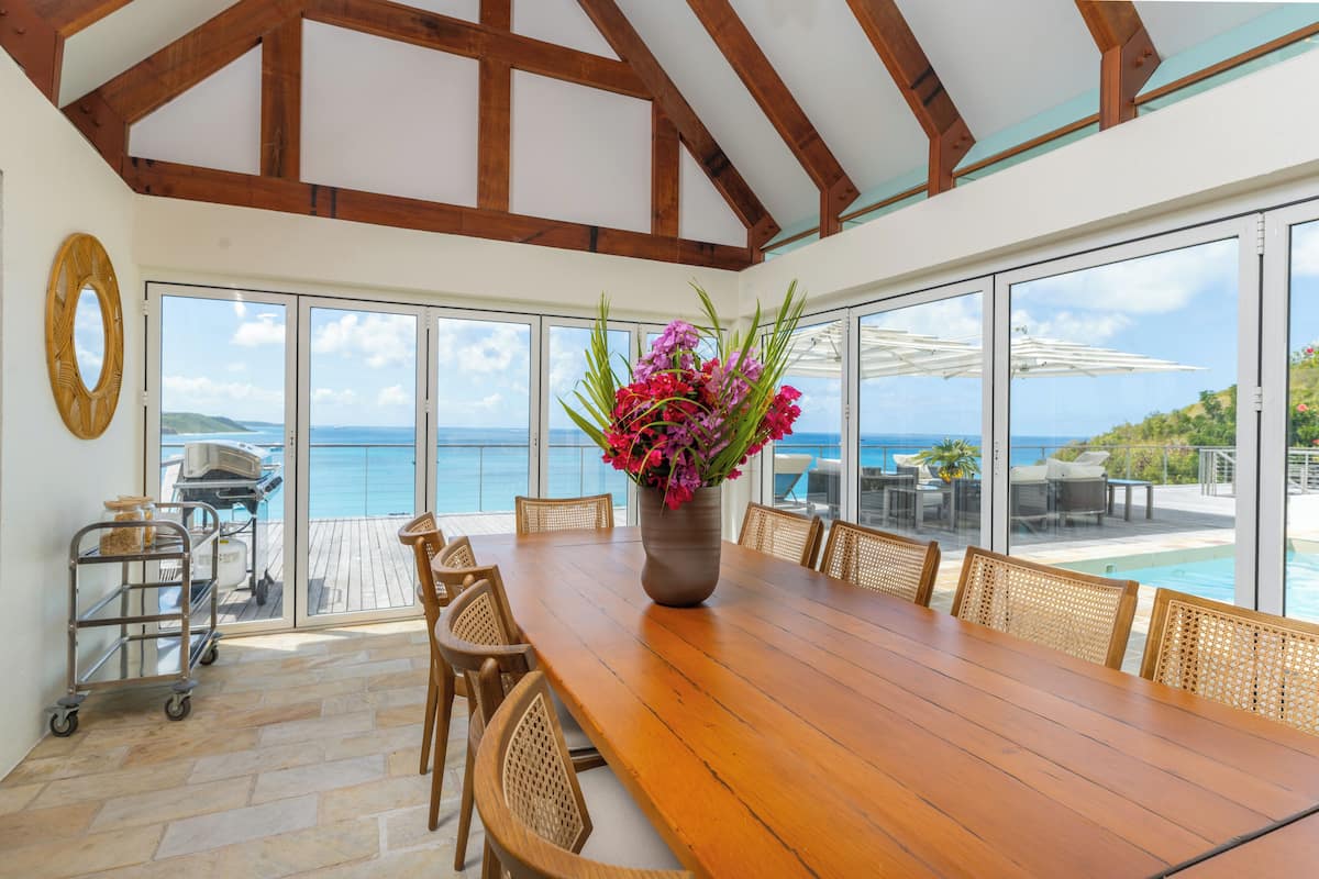 A wooden dining table with wicker chairs, a vase of flowers, and a view of the ocean through large windows.