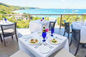 A dining area with tables set for two, overlooking a coastal view with boats.