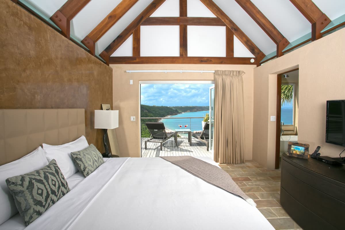 A bedroom with a large bed, a TV, and a view of a balcony with a pool and a scenic landscape.