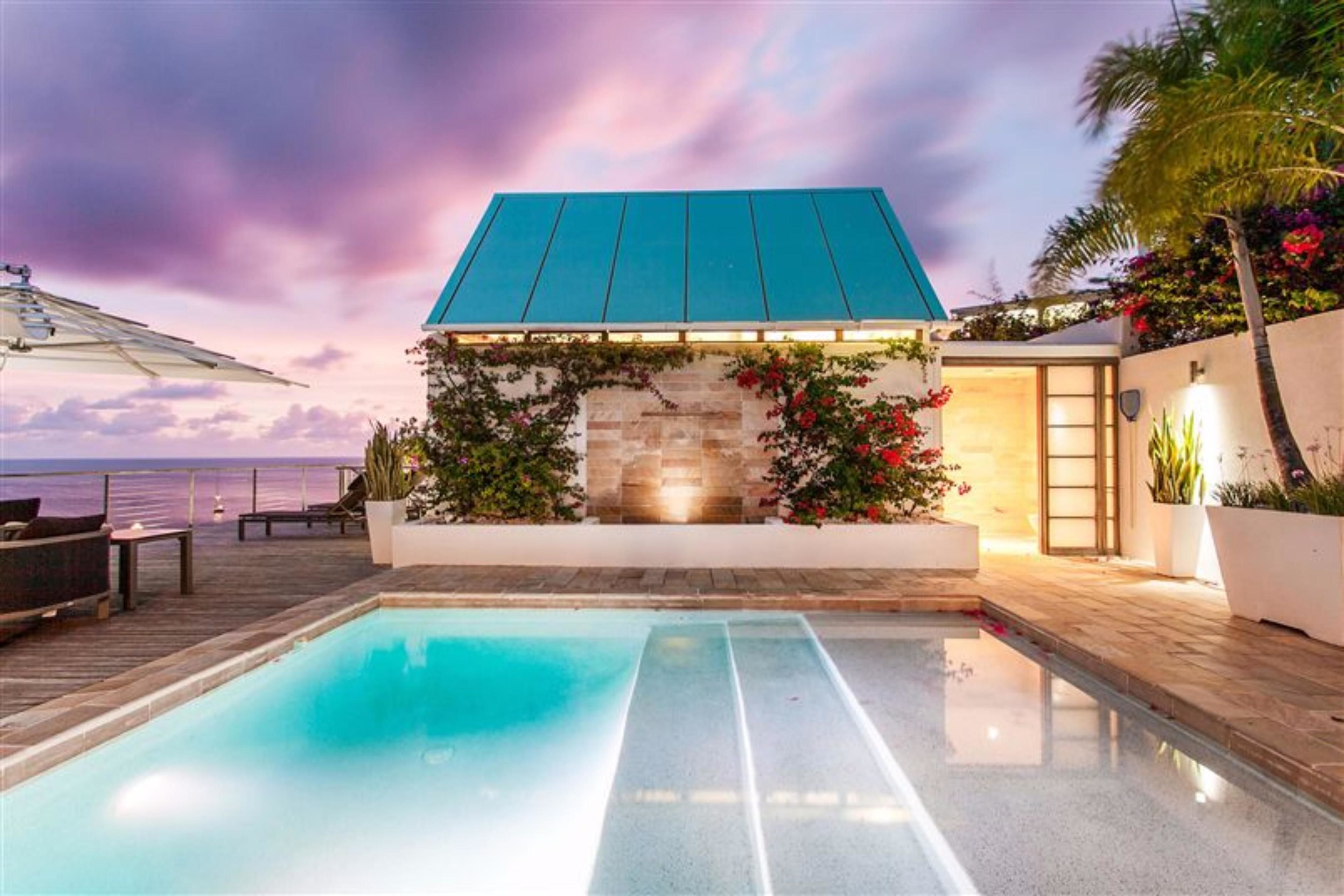 A pool area with a stone wall and a blue roof structure, overlooking the ocean.