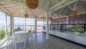 Un espace extérieur abrité avec des places assises et une vue sur la mer, une vitrine réfrigérée où sont présentés les desserts, et une table blanche avec des chaises.