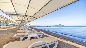 Une plage bordée de rangées de transats sous des parasols, une jetée en arrière-plan et une mer calme.