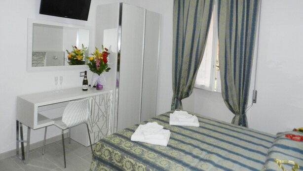A hotel room with a bed, a desk with a chair, a mirror, a TV, and a window with curtains. - Vatican Rome B&B (Rome)