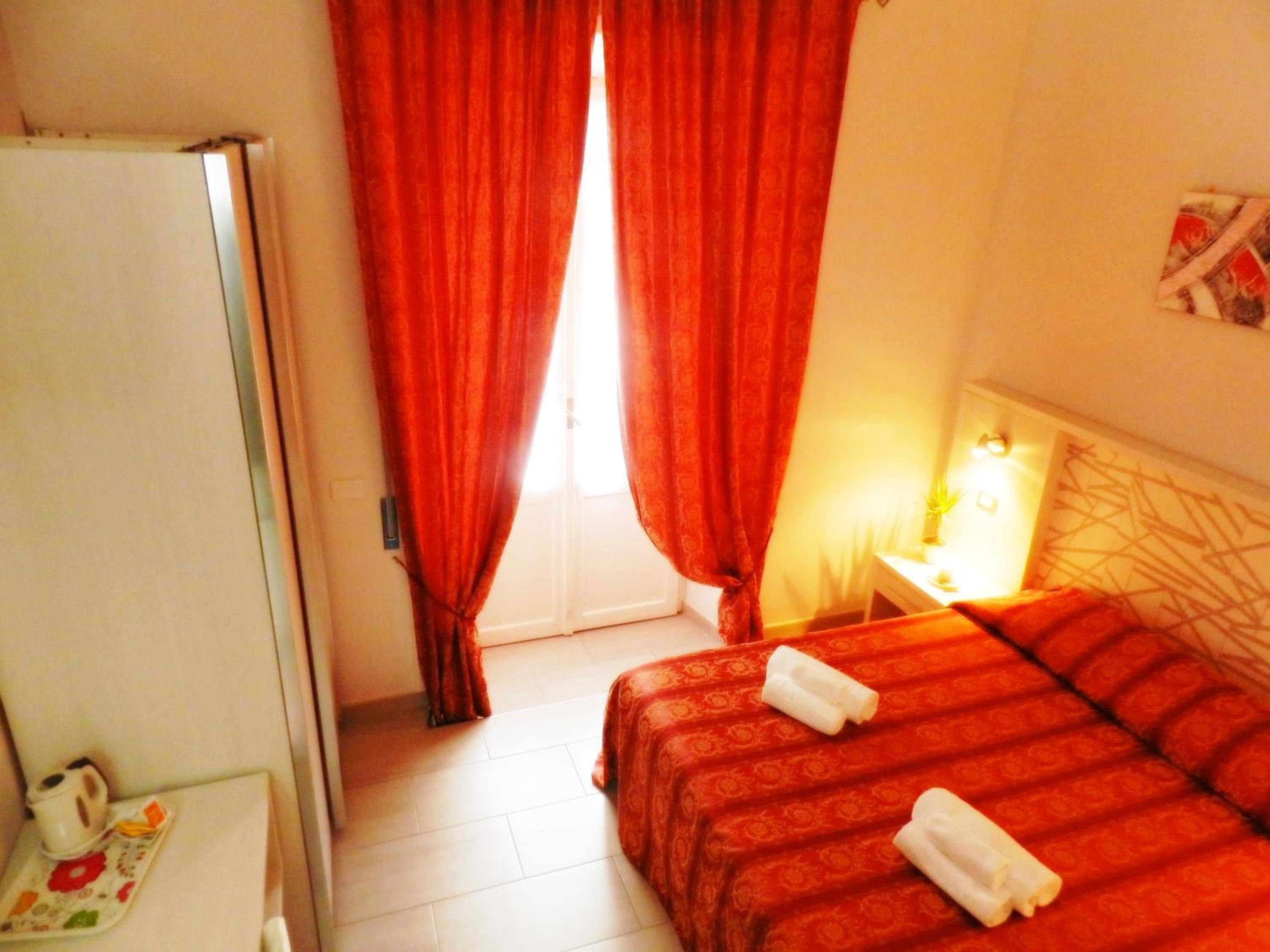 A hotel room with a bed, a nightstand with a lamp, a window with red curtains, and a door.