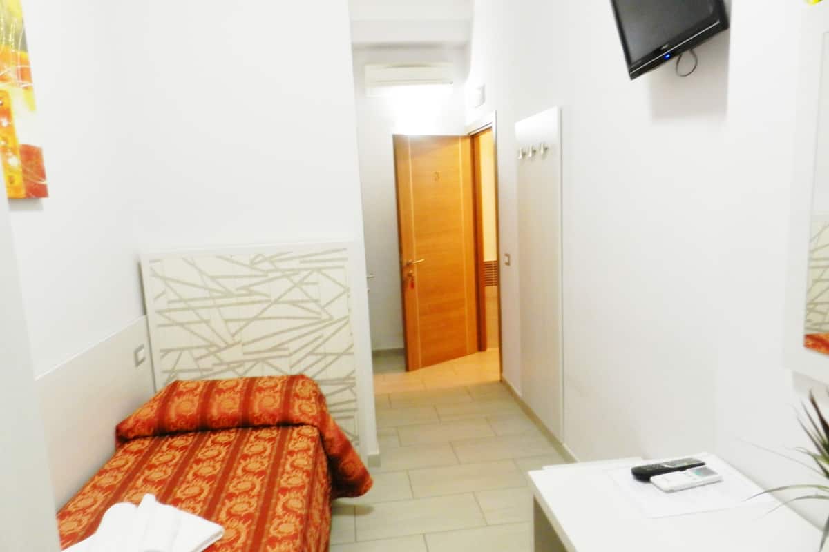 Una habitación de hotel estrecha con una cama, un escritorio, un televisor y una puerta que da a otra habitación.