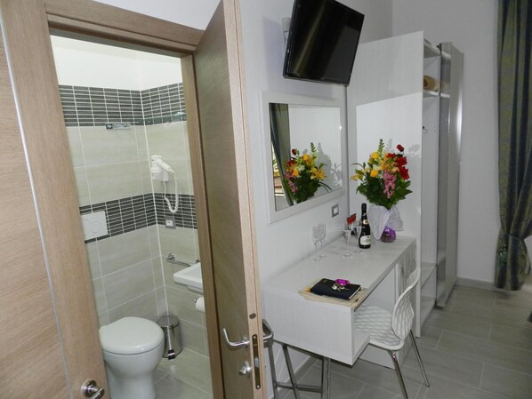 A modern bathroom with a toilet, shower, and a vanity with a mirror, flowers, and a TV.