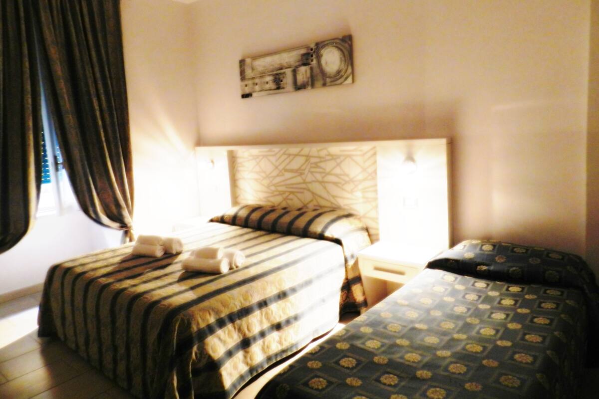 Habitación de hotel con dos camas, un ventanal con cortinas, un reloj de pared y una mesita de noche.