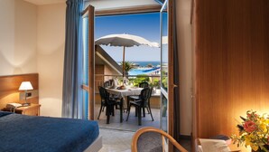 A hotel room with a bed, a desk with a lamp, a chair, a table with chairs, and a balcony with a table, chairs, and an umbrella. - Continental Resort (Pisa)