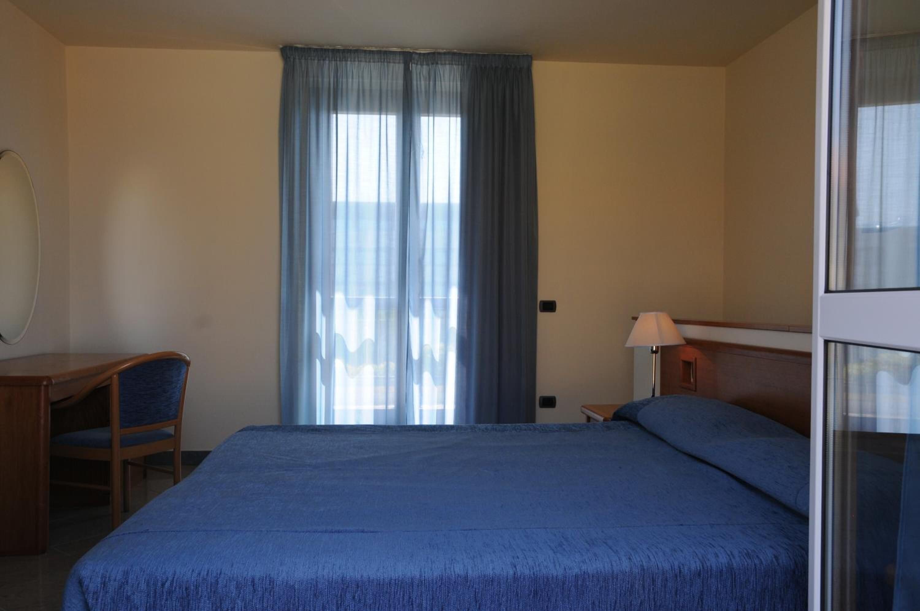 A hotel room with a bed, a desk, a chair, a lamp, and a window with curtains.