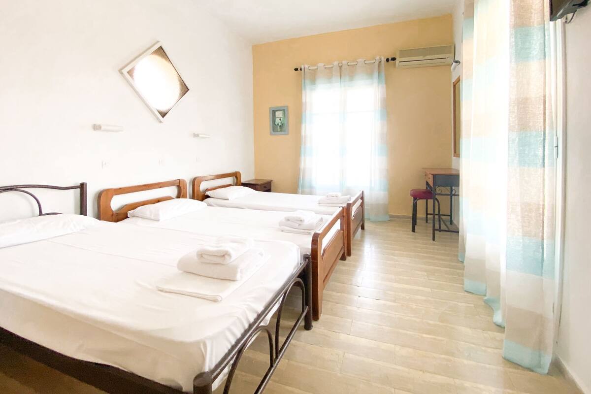 A hotel room with three single beds, a small desk, a chair, and an air conditioning unit.
