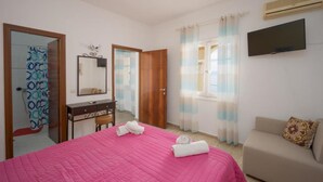 A bedroom with a pink bed, a wooden wardrobe, a desk with a mirror, a TV on the wall, and a sofa. - El Greco Hotel by Estia (Corfu)