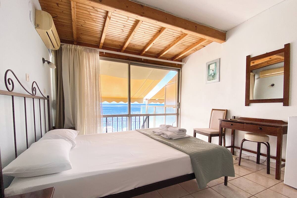 A hotel room with a bed, a desk, a chair, a window with a view of the sea, and a ceiling with wooden beams.