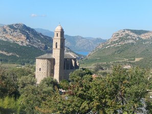 A stone church with a bell tower, surrounded by lush greenery and mountains in the background. - Hôtel La Palma (Patrimonio)
