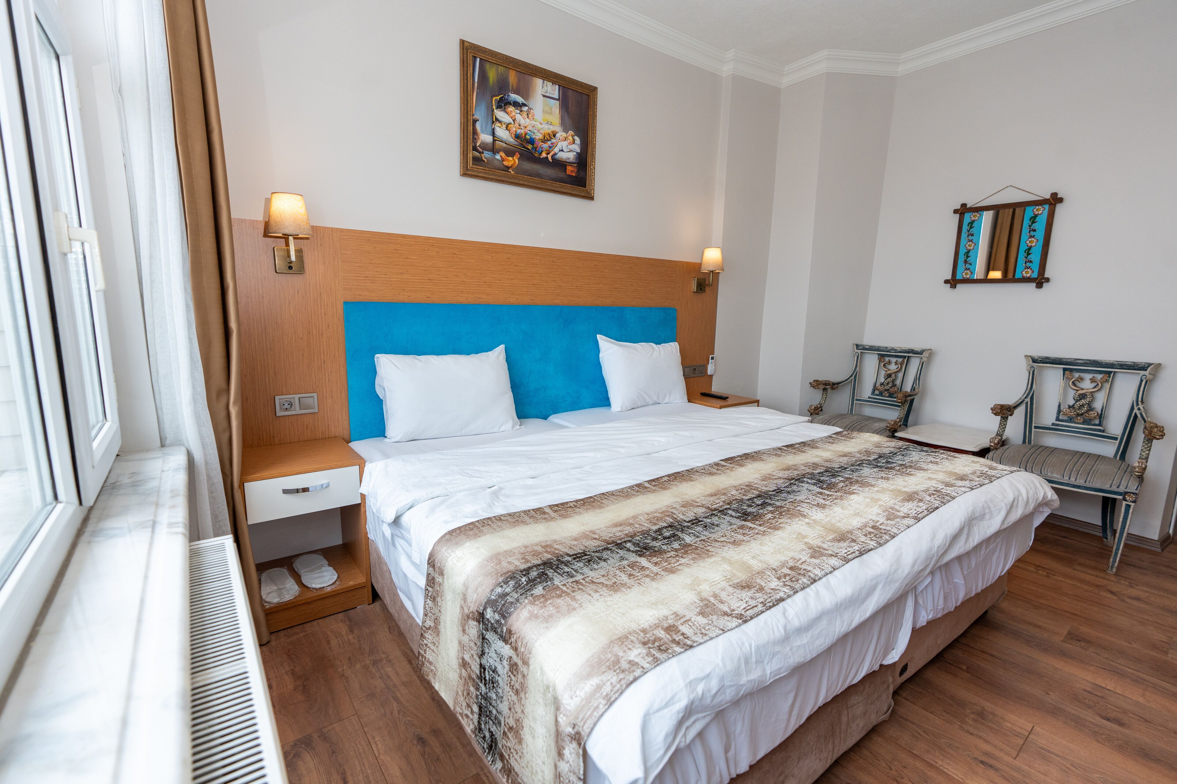A hotel room with a large bed, wooden headboard, two bedside tables, a nightstand, a window with curtains, a wall-mounted painting, and two decorative chairs.