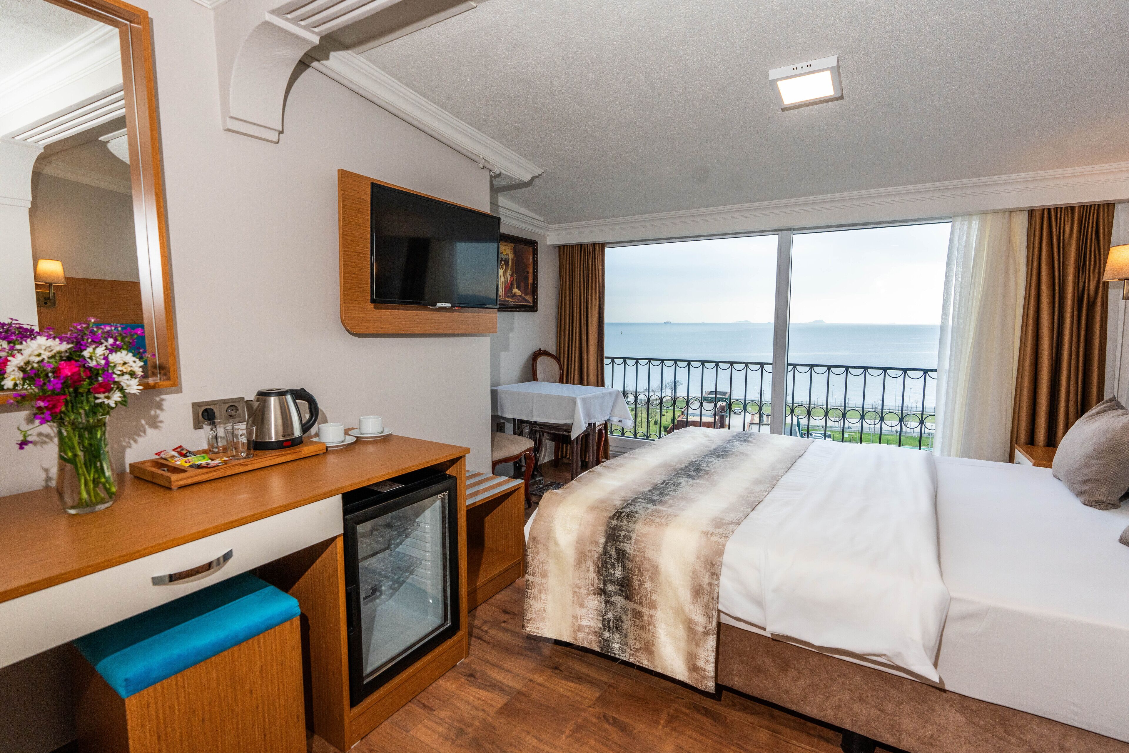 A hotel room with a bed, a desk with a coffee maker, a TV, a small fridge, a window with a view of the sea, and a balcony with a table and chairs.