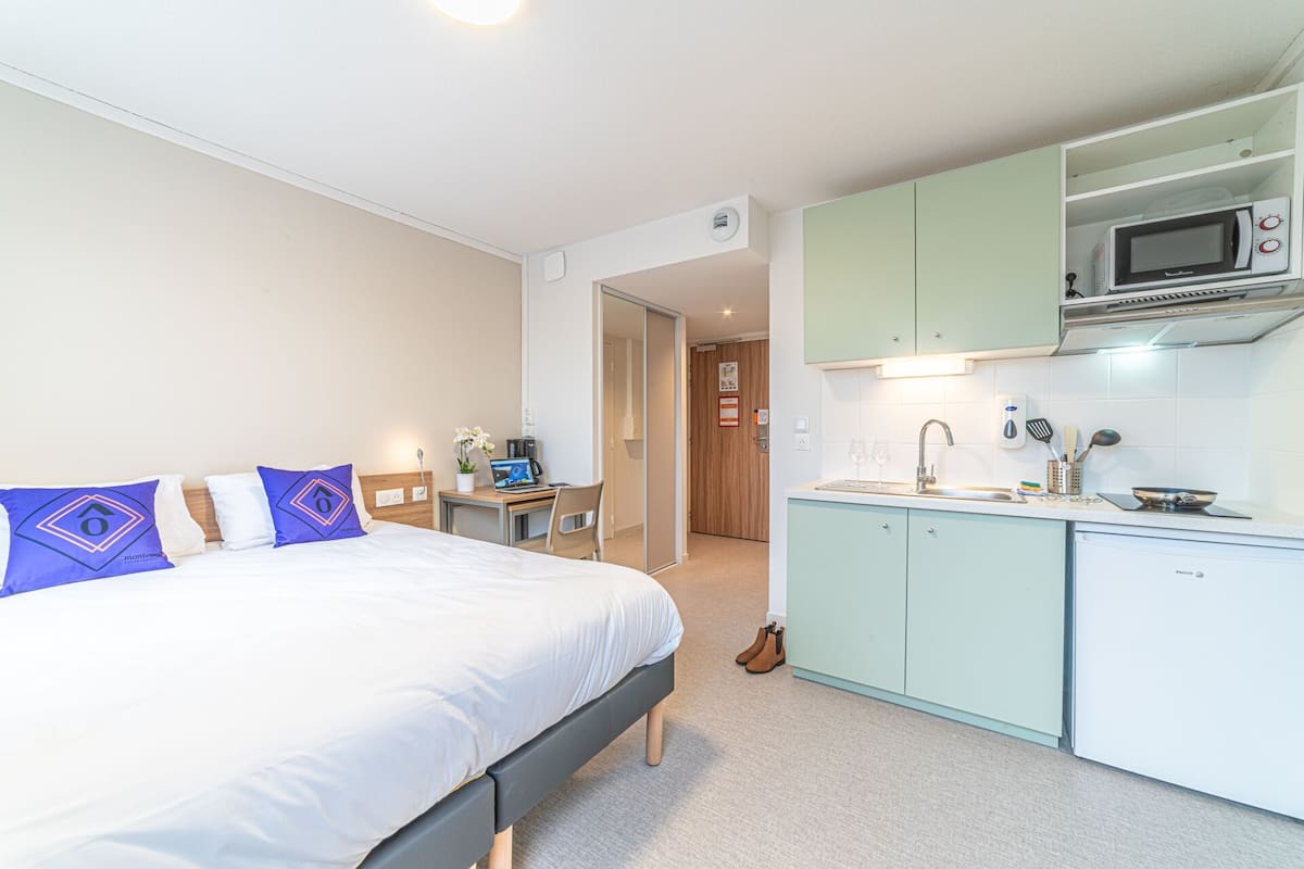 A compact hotel room with a bed, a small desk with a laptop, a kitchenette, and a door leading to another room.