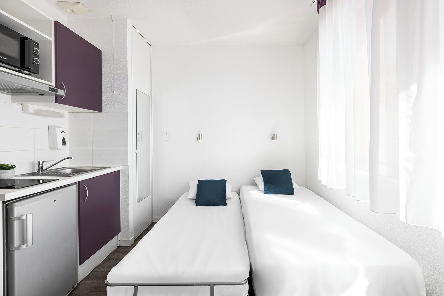 A compact hotel room with a kitchenette, a bed with two pillows, a window with sheer curtains, and a white wall.