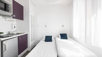 A compact hotel room with a kitchenette, a bed with two pillows, a window with sheer curtains, and a white wall.