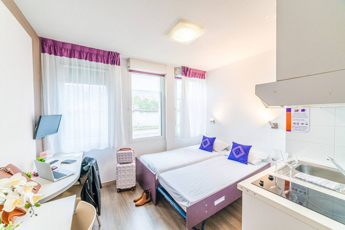 A hotel room with a bed, a desk with a laptop, a suitcase, and a kitchenette.