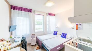 A hotel room with a bed, a desk with a laptop, a suitcase, and a kitchenette.