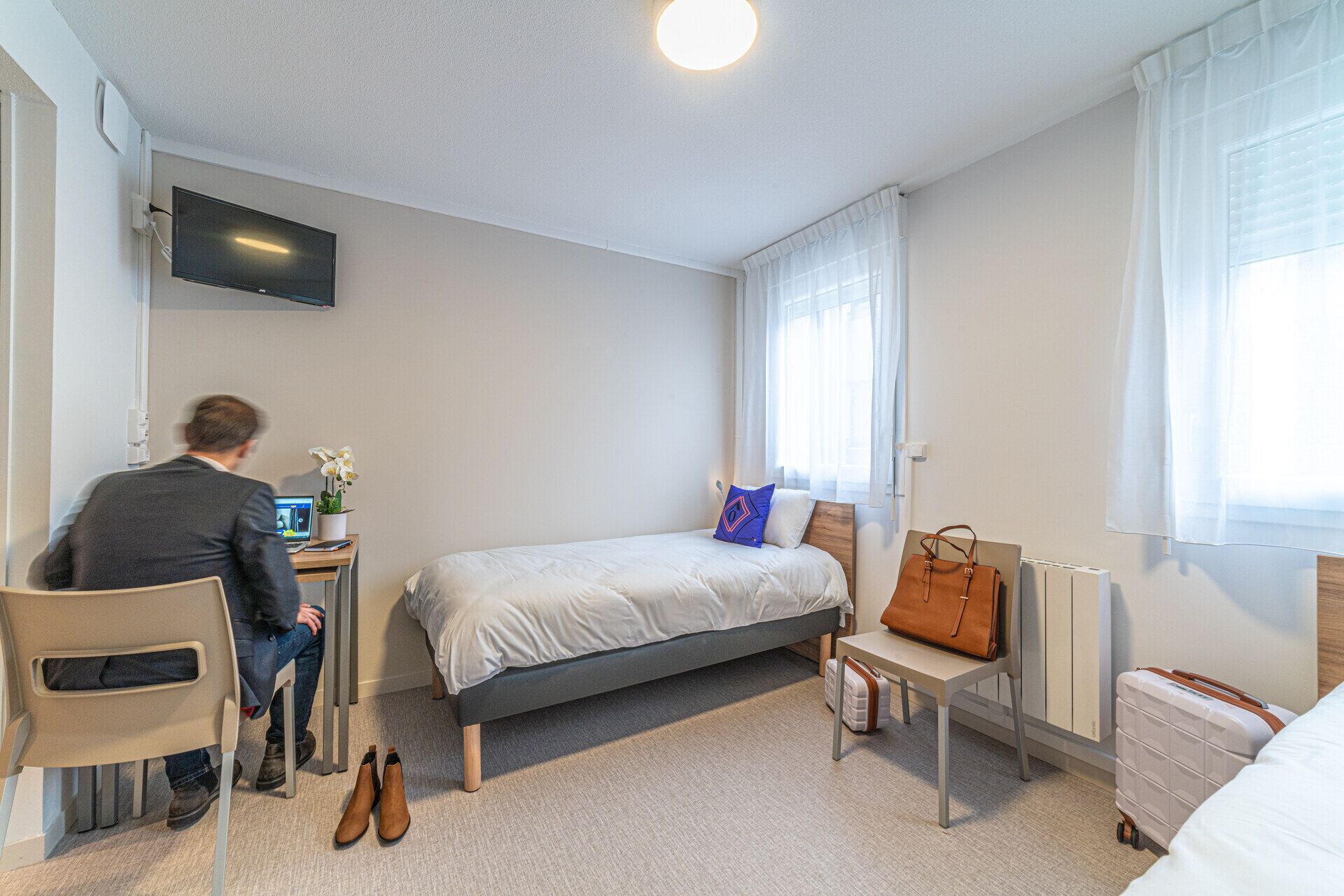 A bedroom with a bed, a desk with a computer, a chair, a radiator, and a suitcase.