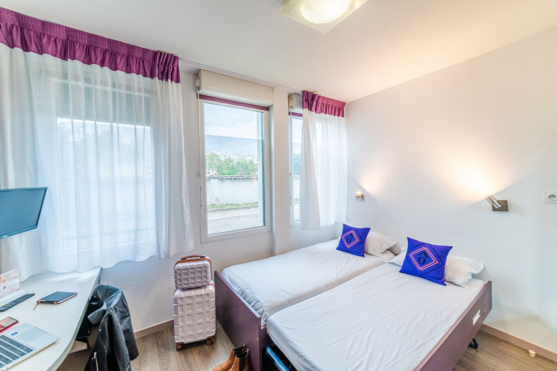 A hotel room with two beds, a desk with a laptop, a suitcase, and a window with curtains.