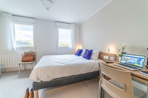 Habitación de hotel con cama, escritorio, laptop, silla, radiador y ventana con cortinas.