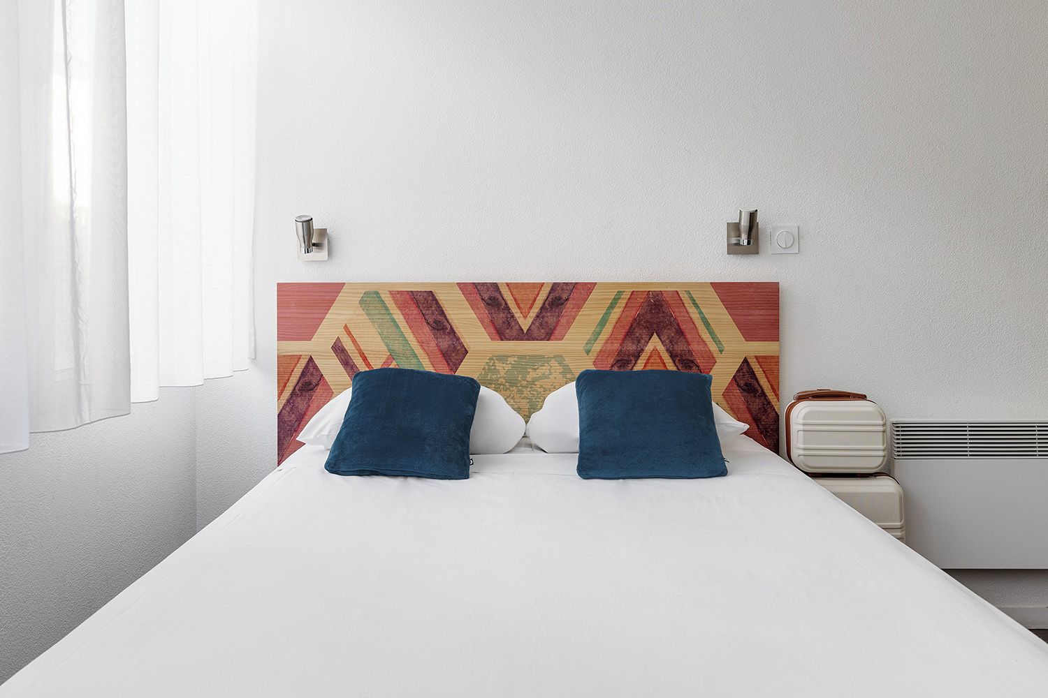 Ein Bett mit zwei Kissen, ein verziertes Kopfteil, ein Nachttisch mit Radio und ein Fenster mit Vorhängen.