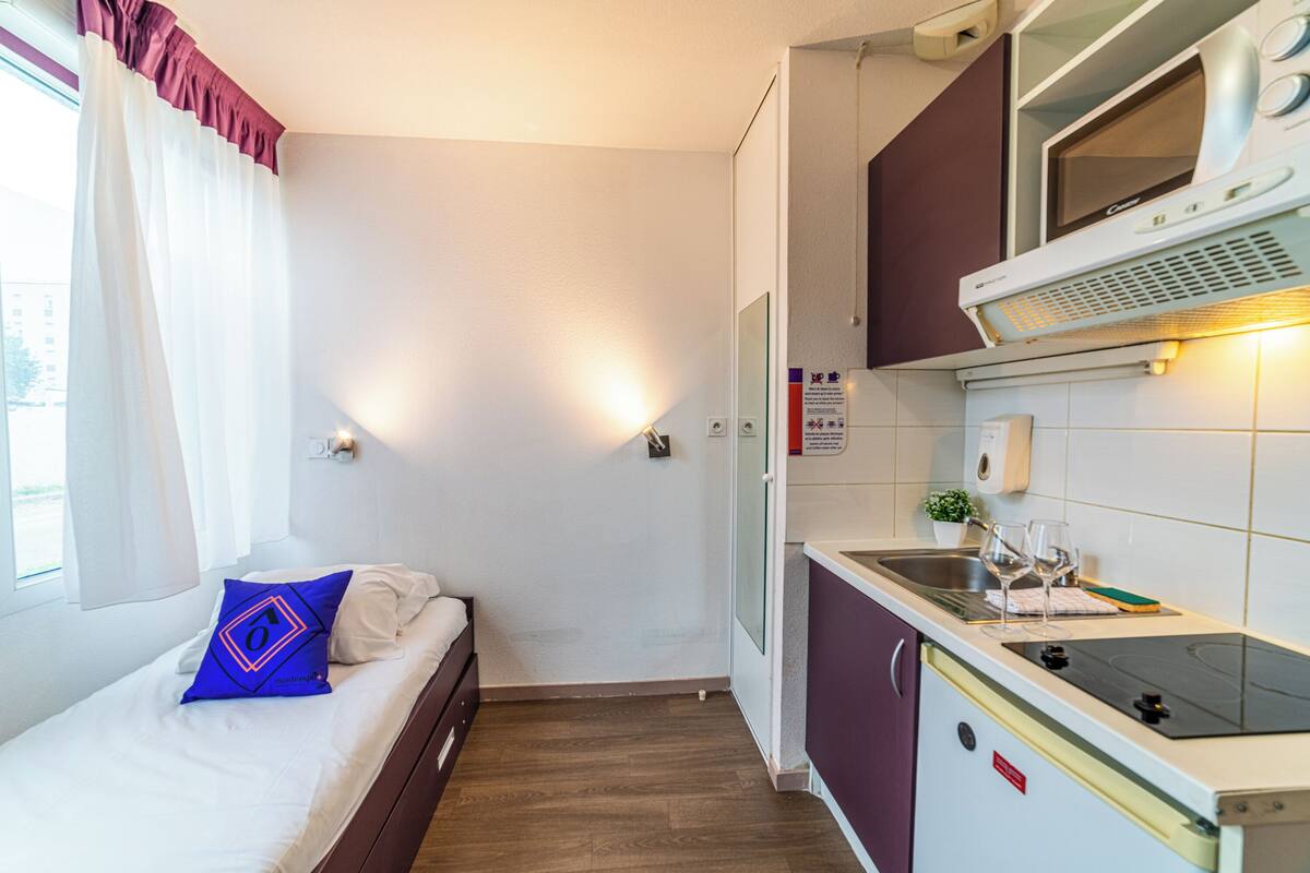 A compact hotel room with a bed, a small kitchenette, and a window with curtains.