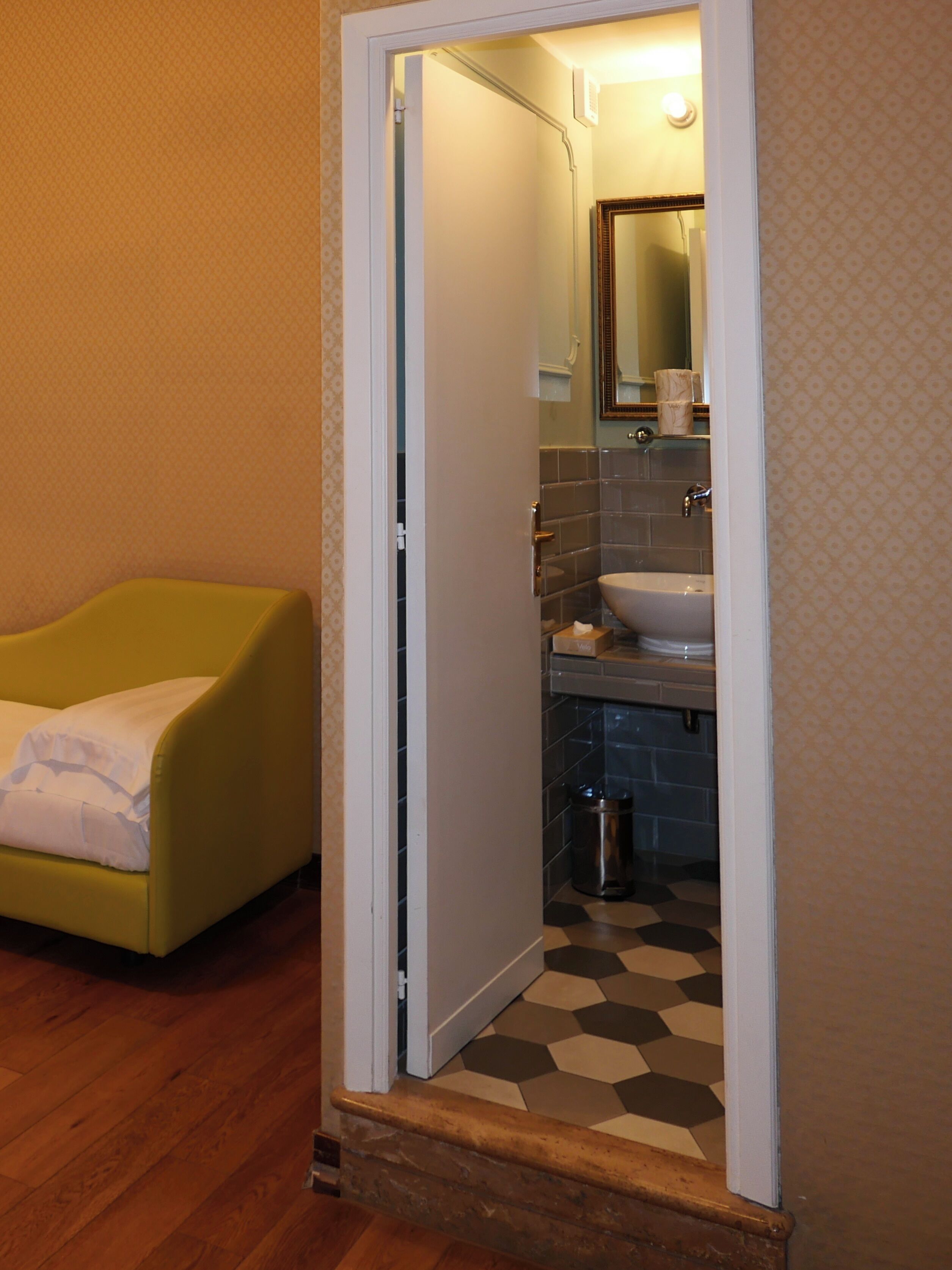 A bathroom with a sink, mirror, and toilet. There is a yellow chair in the adjacent room.