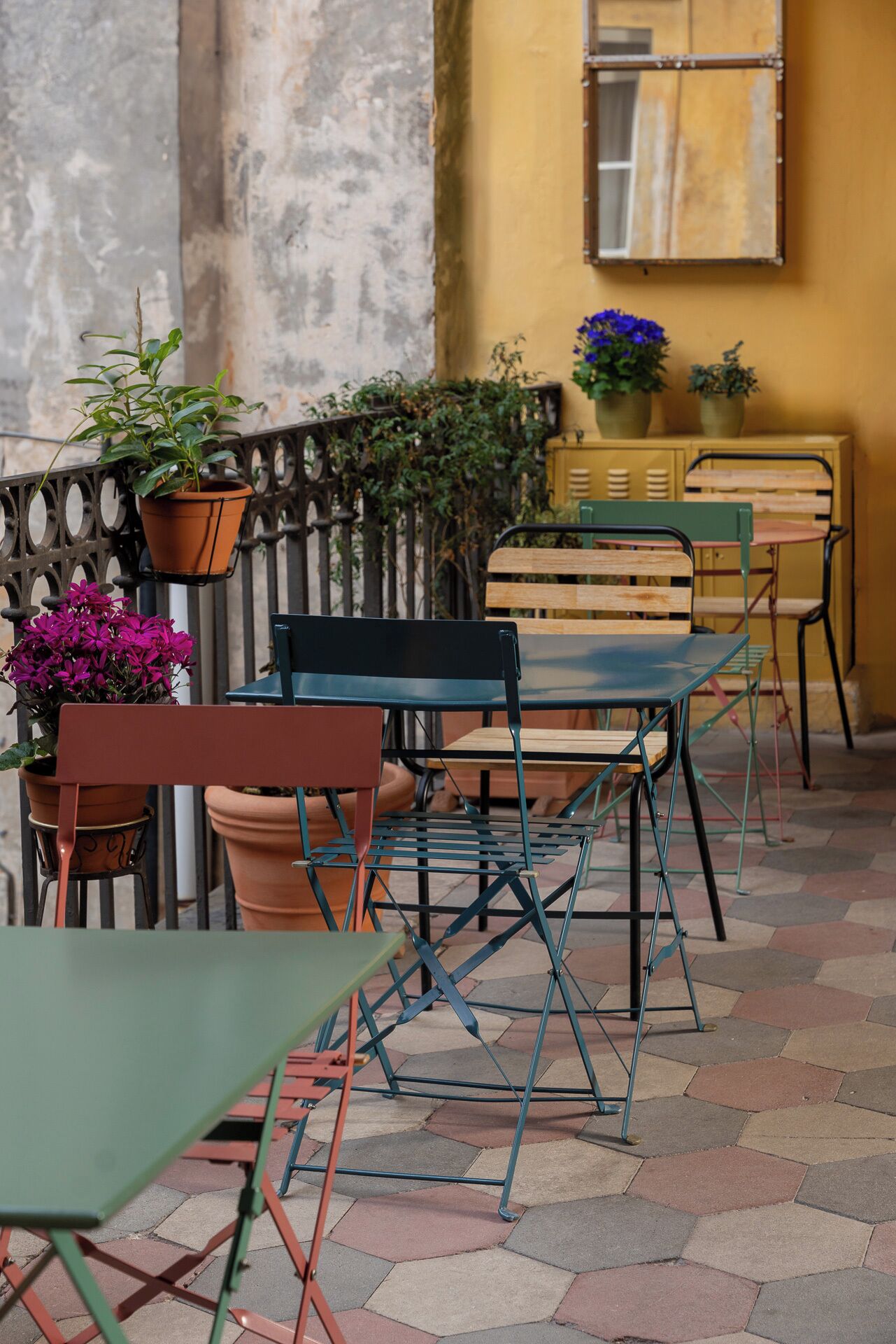An outdoor seating area with metal chairs and tables, potted plants, and a yellow wall in the background.