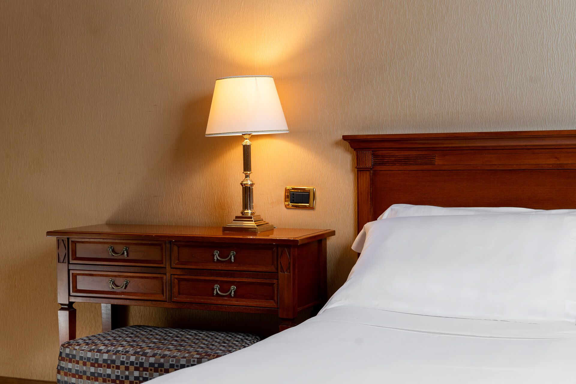 A hotel room with a wooden nightstand, a lamp, a bed with white linens, and a patterned ottoman.