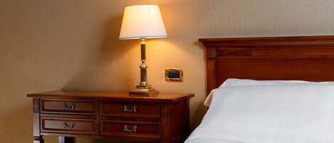 A hotel room with a wooden nightstand, a lamp, a bed with white linens, and a patterned ottoman.