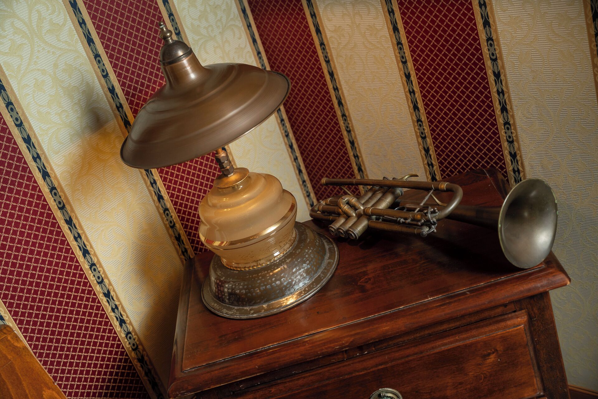 A vintage desk lamp with a brass base and a glass shade, placed on a wooden desk with a drawer.