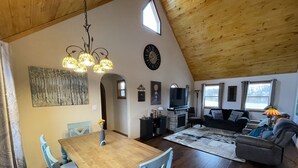 Un amplio espacio de estar con techo de madera, una mesa de comedor con sillas, una chimenea, un televisor y un sofá.