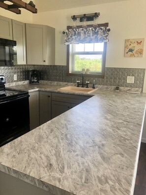 A kitchen with a marble countertop, a built-in stove, a coffee maker, and a window with a floral curtain.