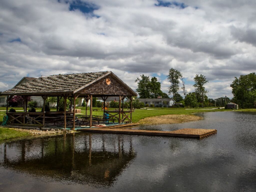 Un porxo al costat d'un llac tranquil amb una terrassa de fusta, envoltat de vegetació i un cel nuvolós.