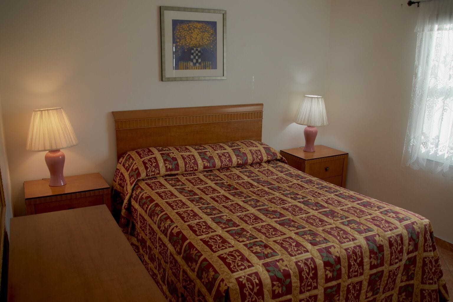 Ein Schlafzimmer mit einem hölzernen Bett, zwei Nachttischen mit Lampen, einem gerahmten Bild an der Wand und einem Fenster mit transparenten Vorhängen.