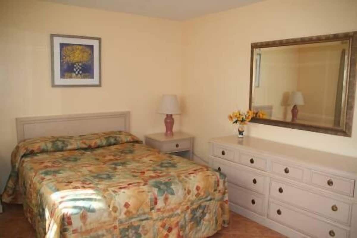 A bedroom with a bed, a nightstand, a dresser, a mirror, and a framed picture on the wall.