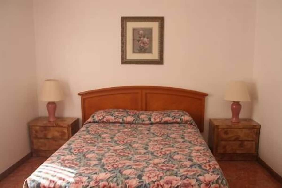 A bedroom with a floral bedspread, wooden headboard, two bedside tables with lamps, and a framed picture on the wall.