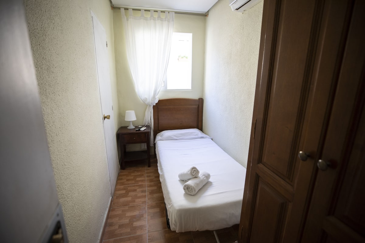 A small, narrow room with a single bed, a nightstand with a lamp, and a window with a sheer curtain.