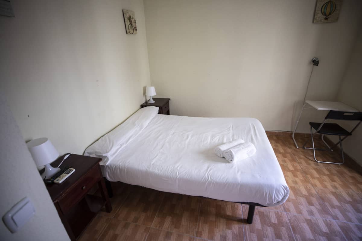 A small, clean room with a single bed, a nightstand with a lamp, a chair, and a desk.