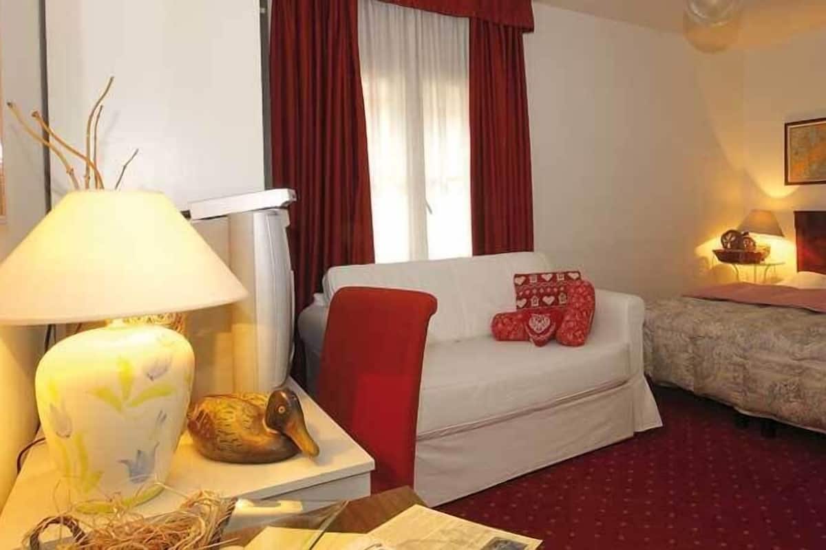 Quarto de hotel com uma cama, uma mesinha de cabeceira com uma luminária, um vaso decorativo e um travesseiro vermelho.