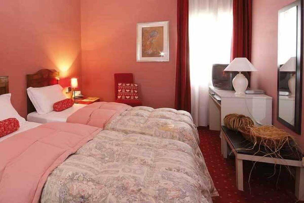Quarto de hotel com duas camas, cada uma com cabeceira de madeira, mesa de cabeceira com abajur e uma pequena mesa com cesta.