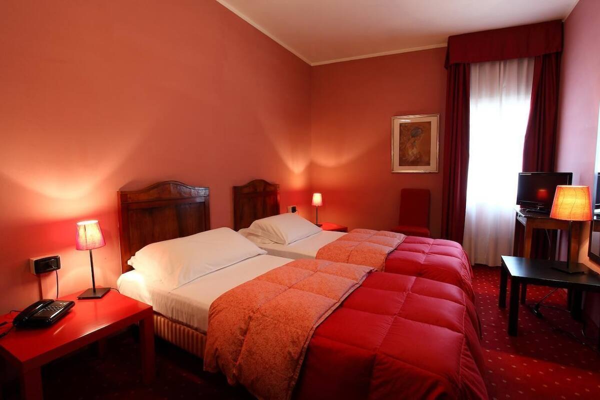 Quarto de hotel com duas camas, televisão, escrivaninha com luminária e um quadro na parede.