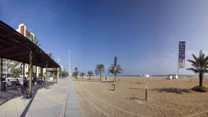 Eine Uferpromenade mit überdachter Sitzgelegenheit, Palmen und einem Sandstrand.